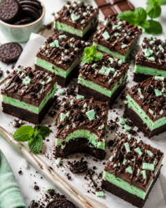 Stacked three-layer chocolate grasshopper fudge with a mint-green center, chocolate drizzle, cookie crumbs, peppermint pieces, and fresh mint.